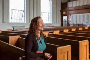 A white woman with long curly red hair wearing a turquoise top and dark cardigan, sitting in semi-profile in a pew of an empty church. She is smiling.