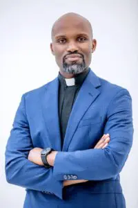 A black man with a bald head, salt-and-pepper goatee wearing a blue blazer and black shirt with clerical collar, standing with his arms crossed in front of a white background