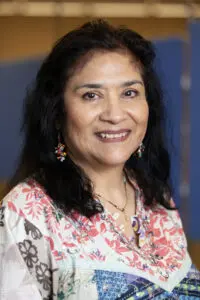 Middle aged woman with brown skin and black shoulder length hair, wearing a multicoloured top. She is smiling.