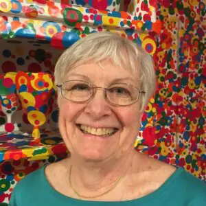 The head and shoulders of a smiling woman with short grey hair and glasses in front of a multicoloured background